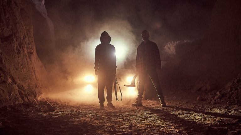 image of two hooded figures backlit by car headlights. one carries a boomerang the other a shotgun.