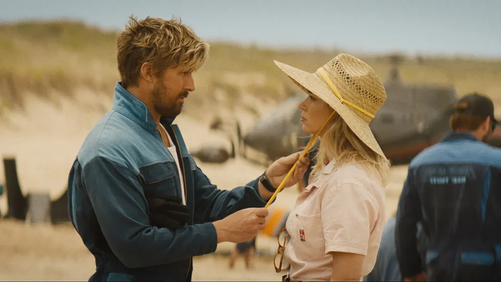 A blonde man (Ryan Gosling) pulls on the toggles of a blonde woman's (Emily Blunt) sun hat, looking deep into her eyes. they are on the sandy, beach set of a film in production.